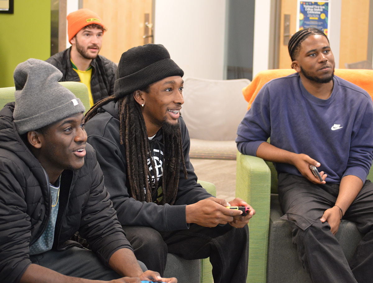 Four students in a lounge on green furniture, two are playing video games and two are watching