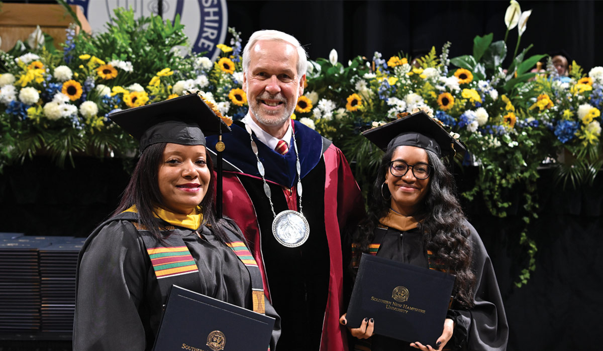Man with white hair in black and red collegiate robes and a medal around his neck, taking a picture with two female students holding diplomas, flowers in the background 