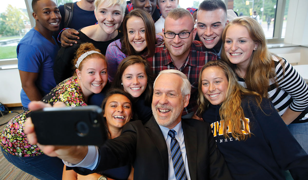 Man with white hair, taking a selfie with 13 students behind him