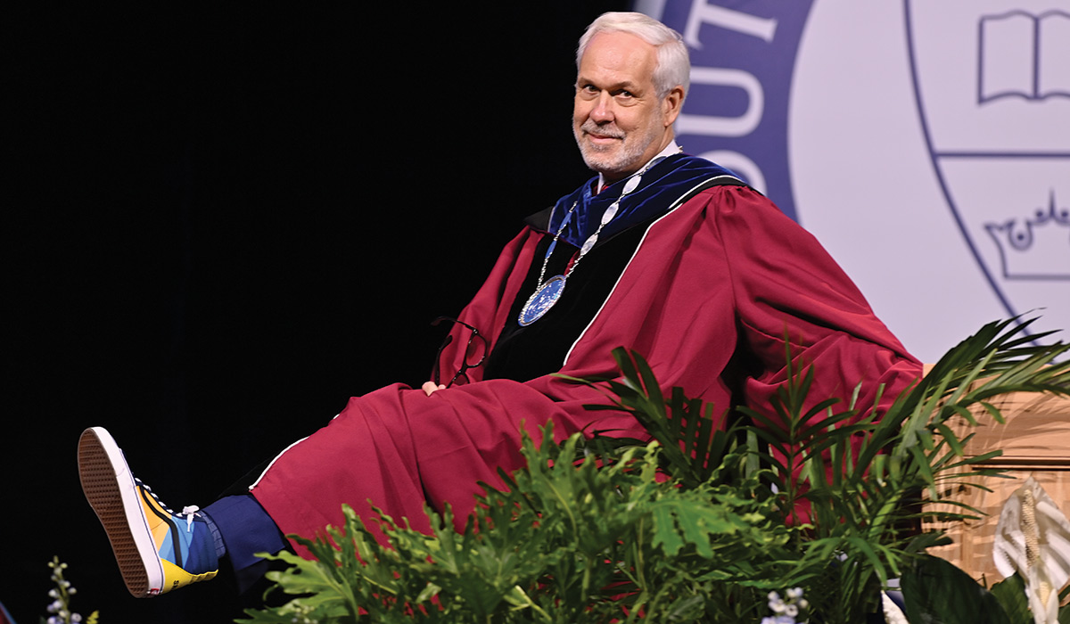 Man in red collegiate robes with one leg raised out in front of him, smiling at camera