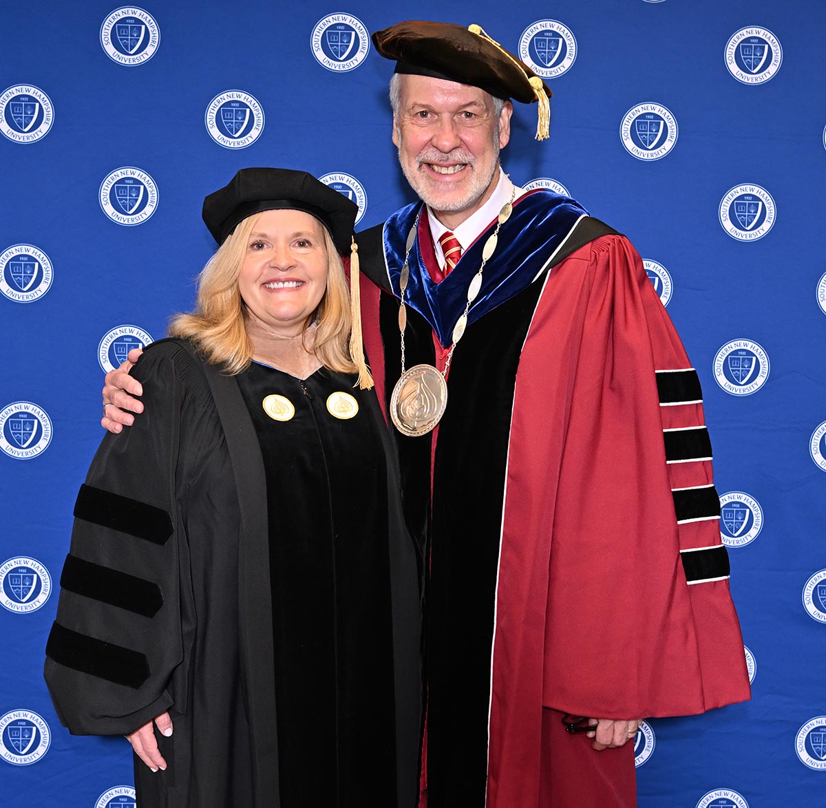 Man in red collegiate robes, a hat with a tassel,  and a medal around his neck, standing next to a woman in black collegiate robes and a hat with a tassel in front of a blue background