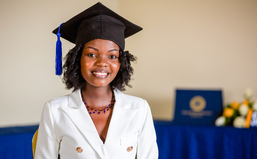 Alexia Umugwaneza ’23 wearing her black graduation cap with a white blazer with gold buttons while standing in front of a table display with a blue table cloth and flowers and diploma sitting on it