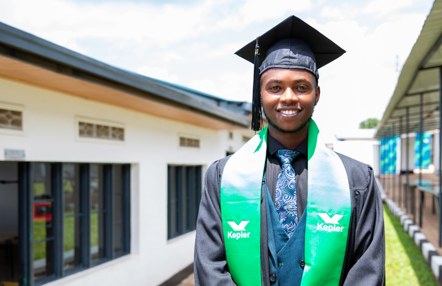 Mupenzi Niyomugabo ’23 standing outside in between two rows of classrooms wearing his black graduation cap, a black suit with a blue tie, and a green stole around his shoulders