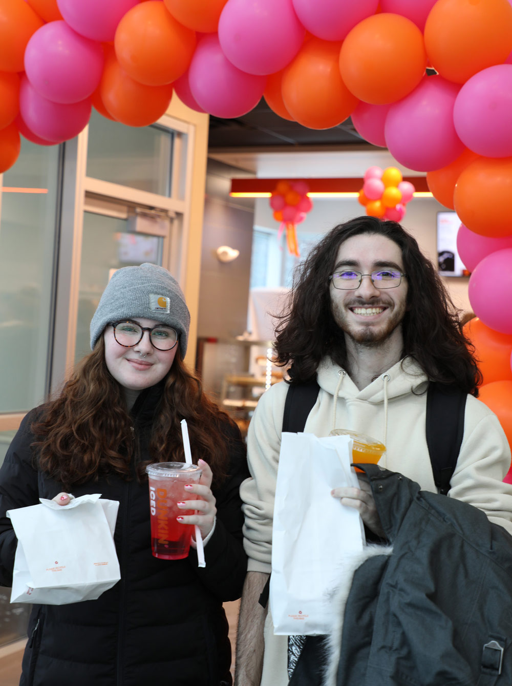 two students with drinks and bags in front of an orange and pink balloon arch in front of a Dunkin'
