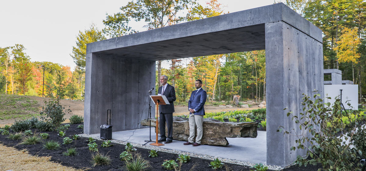 man giving a speech at a podium with another man behind him at the Labyrinth Memorial