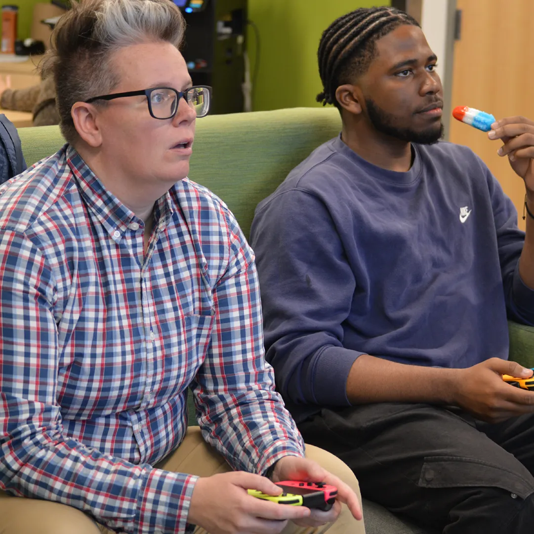 Two people playing video games looking at screen in distance while one eats a popsicle