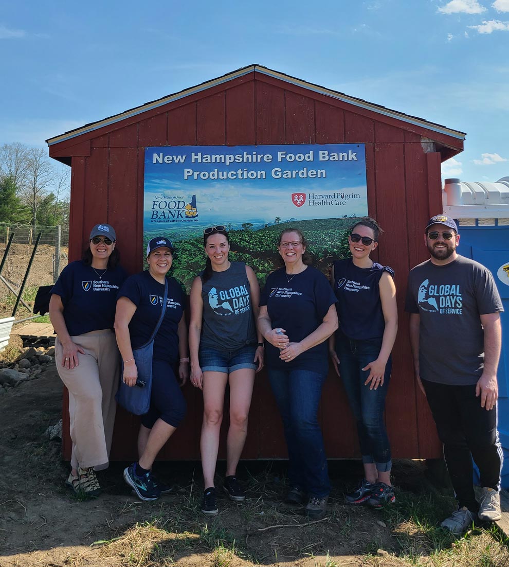 volunteers wearing SNHU and Global Days of Service t-shirts smile while standing in front of a shed that holds a sign that reads: New Hampshire Food Bank Production Garden