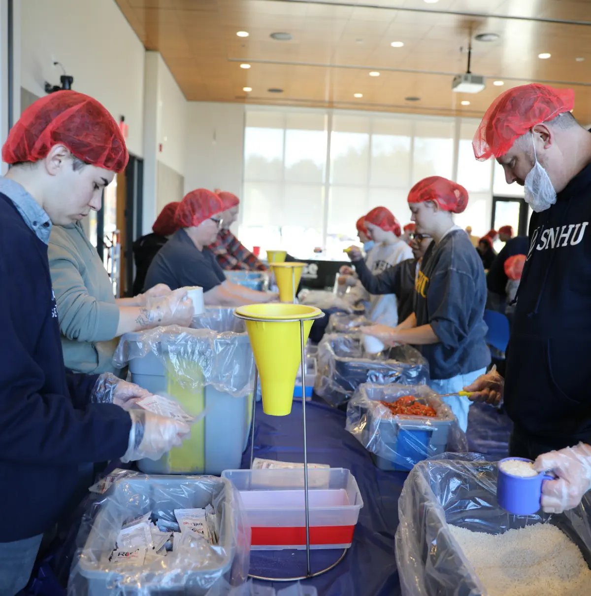 Portrait orientation indoor photograph close-up view of a long table full of volunteers packing meals for a Rise Against Hunger service project