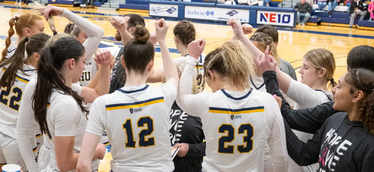 Landscape orientation indoor photograph close-up view action shot of the SNHU woman's basketball team at the end of a court side team huddle all putting their fists together in the air