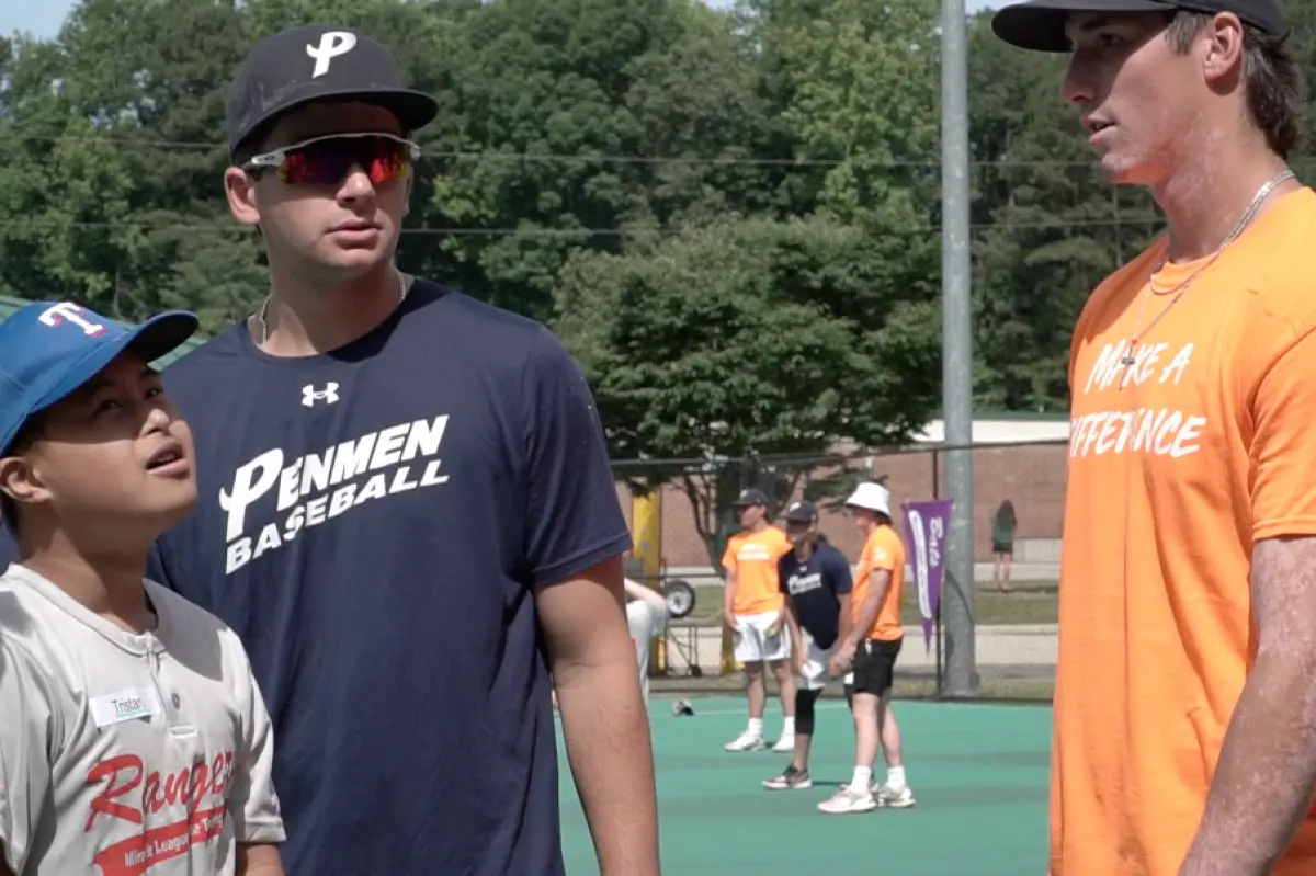 Portrait orientation outdoor photograph close-up view of two SNHU baseball team players and a young boy all gathered nearby each other at a special exhibition game alongside children from The Miracle League of the Triangle