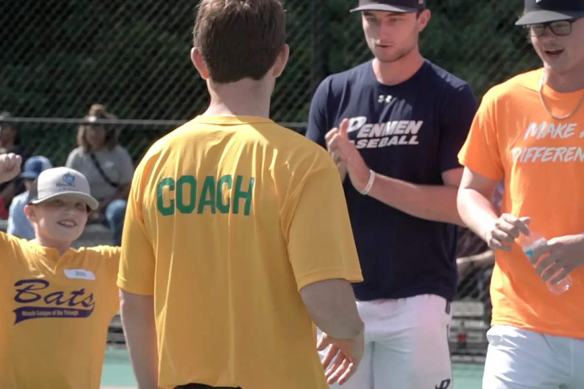 Portrait orientation outdoor photograph close-up view of a young boy holding his hands up in celebration while approaching a coach as there are two SNHU baseball team players looking upon the little young boy to congratulate him during a special exhibition game alongside children from The Miracle League of the Triangle