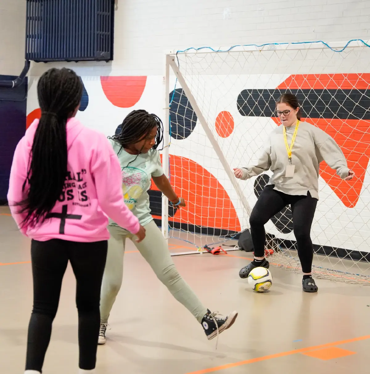 Portrait orientation close-up indoor photograph view action shot of a service-learning student from The Chandler Center playing soccer with two middle schoolers at the Amiko Youth Program, which offers after-school programming for newly arrived and first-generation American youth
