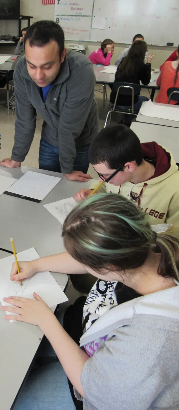A teacher leans over a desk, guiding two students as they work on pencil sketches in a classroom. The students focus on their drawings while other classmates engage in discussions in the background.