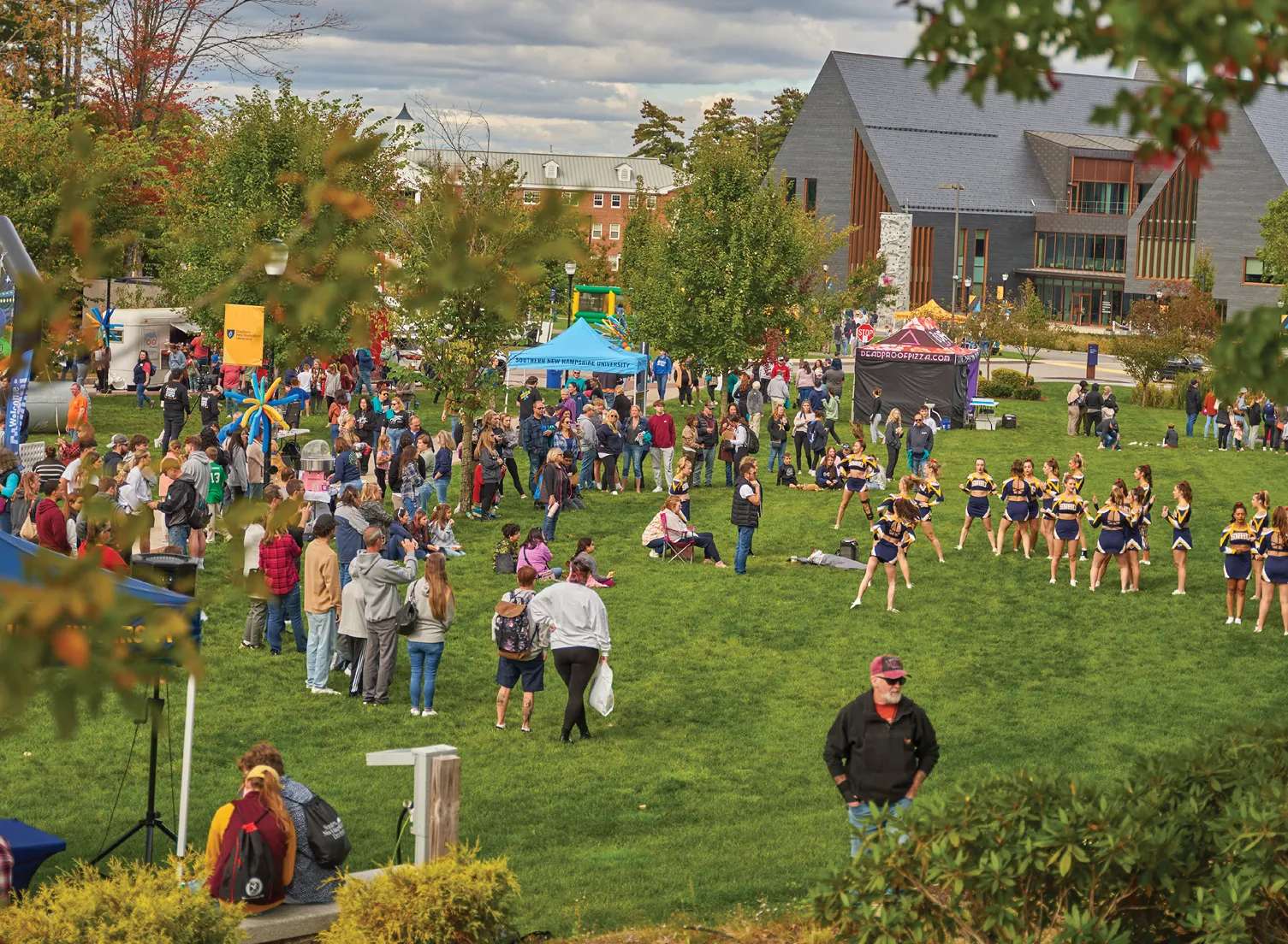 An outdoor campus event with a large crowd gathered on a green lawn. People visit tents and booths while a group of cheerleaders performs. A modern building with large windows stands in the background.