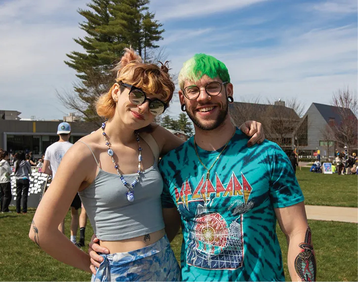 A young couple poses together on a college campus lawn, both smiling. The woman wears sunglasses, a cropped tank top, and a beaded necklace, while the man sports a tie-dye Def Leppard t-shirt and green-dyed hair.