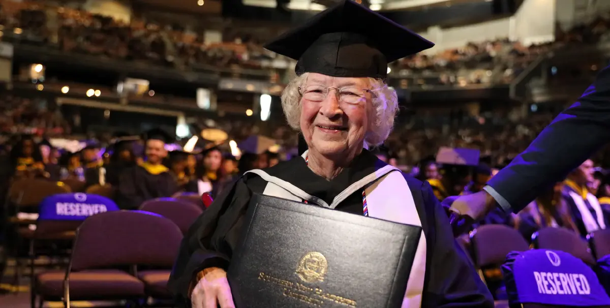 Landscape orientation indoor cropped close-up photograph view of Annette Roberge smiling in her black graduation cap and gown with a white stole around her shoulders while holding her Southern New Hampshire University diploma cover in her hands as she is seated down in a chair