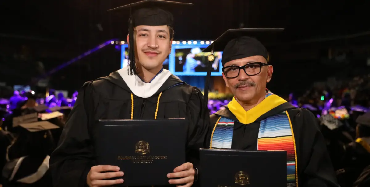 Landscape orientation indoor cropped close-up photograph view of David Perez and his son Angel grinning in their own respective black graduation cap and gown while holding their own respective Southern New Hampshire University diploma cover in their hands as they are standing next to each other