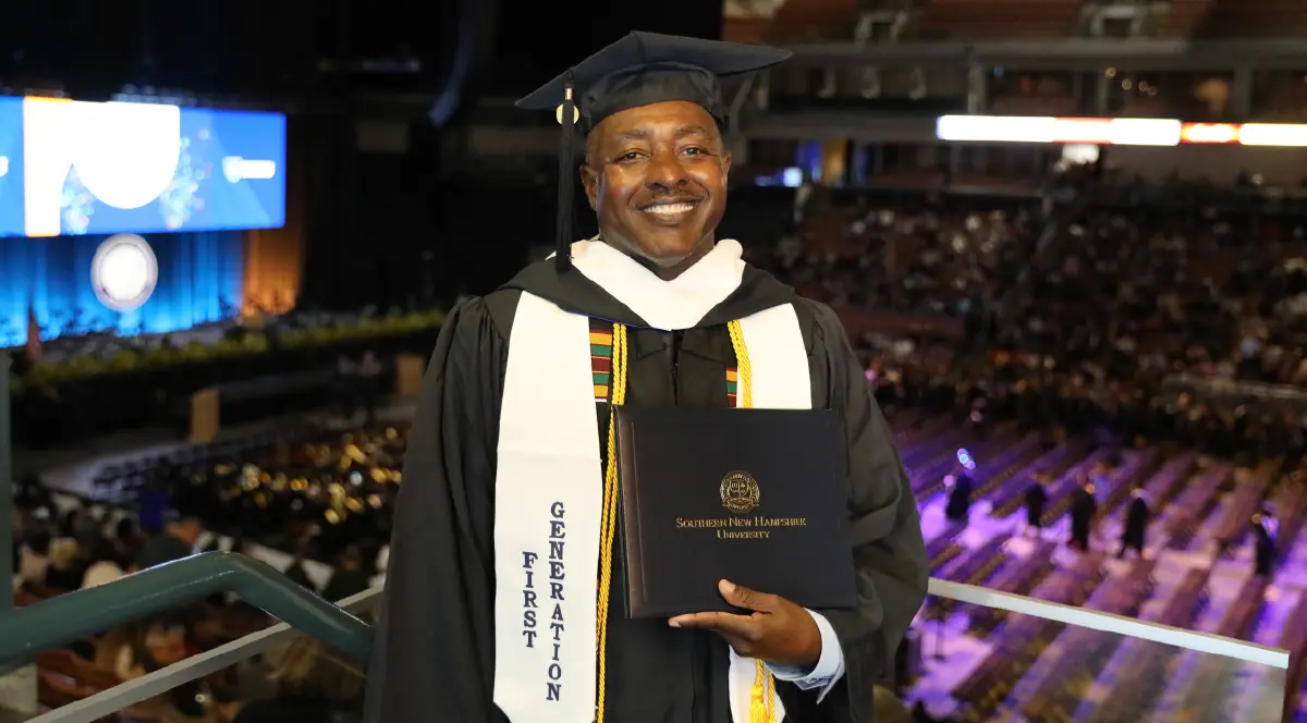 Landscape orientation indoor cropped close-up photograph view of Leon Adams smiling in his black graduation cap and gown with a white stole around his shoulders that says FIRST GENERATION while holding his Southern New Hampshire University diploma cover in his hands as he is standing