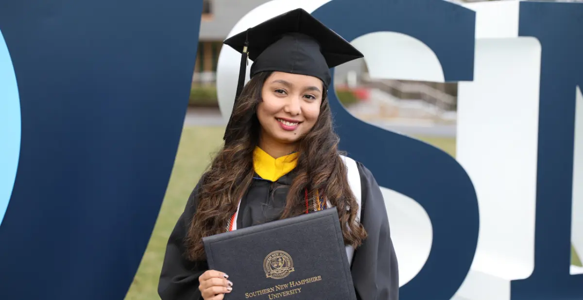 Landscape orientation outdoor cropped close-up photograph view of Riya Thapa smiling in her black graduation cap and gown with a white stole around her shoulders while holding her Southern New Hampshire University diploma cover in her hands as she is standing