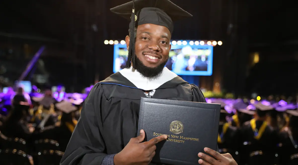 Landscape orientation indoor cropped close-up photograph view of Aaron Mukuna smiling in his black graduation cap and gown while holding and pointing toward his Southern New Hampshire University diploma cover in his hands as he is standing