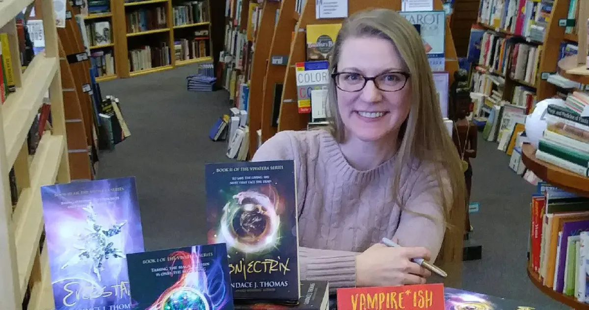 Landscape orientation indoor cropped close-up photograph view of Candance Thomas smiling in a dark beige cardigan sweatshirt plus thin outer frame prescription see through eyeglasses while she is holding a marker in her right hand as she is seated down in a chair behind a table full of some of her novels on display for the public to see within a library setting