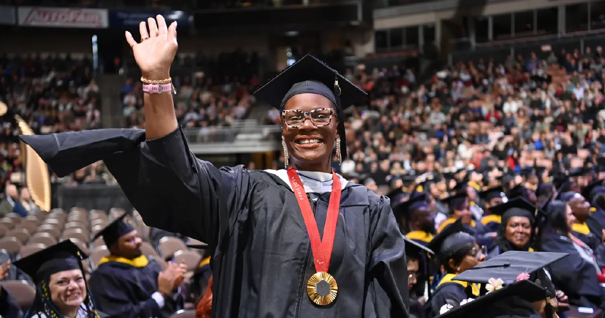 Landscape orientation indoor cropped close-up photograph view of Nicky Nieves smiling in her black graduation cap and gown plus thick outer frame see-through prescription eyeglasses with a gold metal attached via a red lanyard around her neck while she is standing and waving her right hand in air in motion amongst a crowd full of other graduates nearby her seated down in chairs