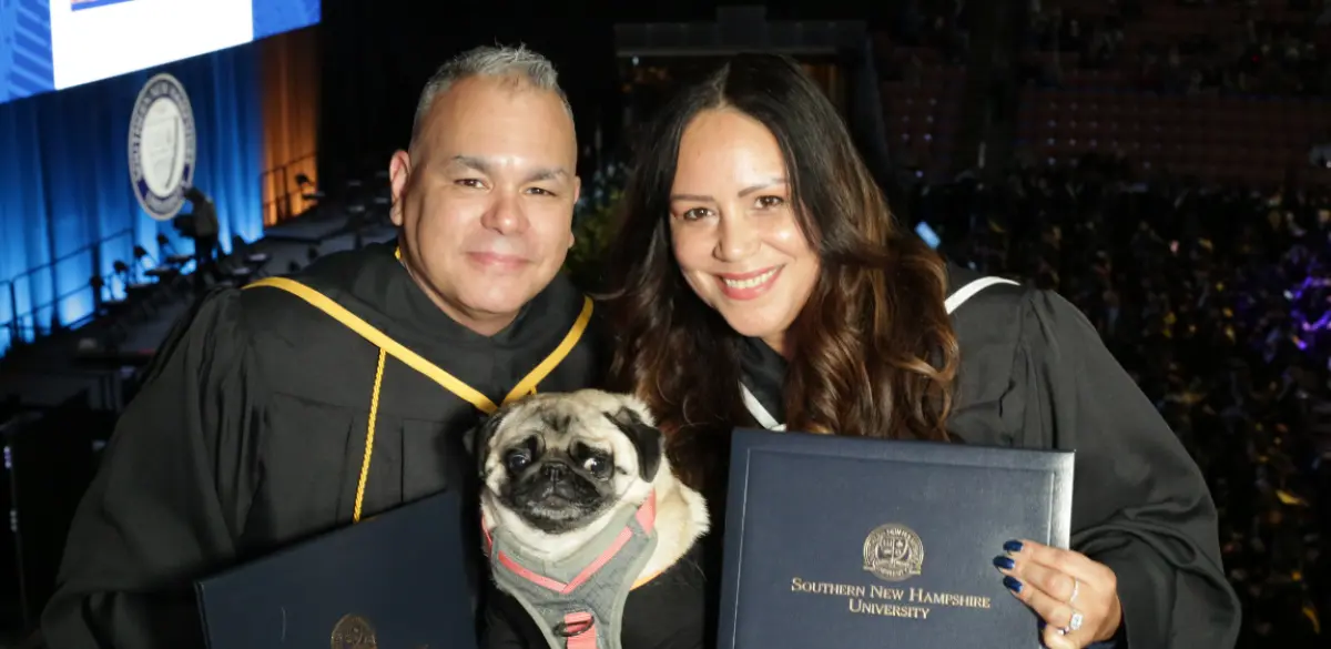 Landscape orientation indoor cropped close-up photograph view of Thedra Lizardi and Robert Vazquez smiling in their own respective graduation gown with their service pug Cora situated in the middle between them as they all are posing for a picture together while Thedra and Robert are holding their own respective Southern New Hampshire University diploma cover in their own hands
