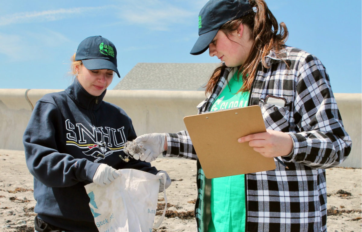 two women wearing gloves and Global Days of Service hats examining a piece of trash found on the beach