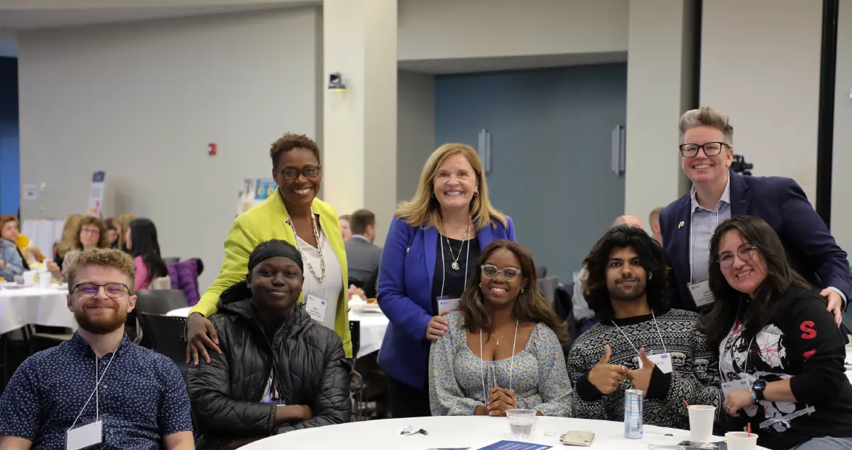 Landscape orientation close-up indoor photograph view of Urban College of Boston President Dr. Yves Salomon-Fernández, President Ryerson, and Dr. Lynn Zlotkowski connecting with student panelists as everyone is gathered together at a table smiling and posing for a picture