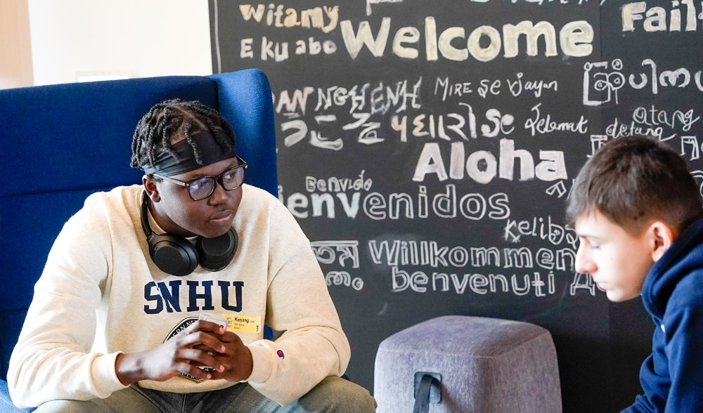 Two young men seated in front of a chalkboard wall with "Welcome" in various languages.