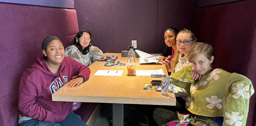 Five people sitting at a table in a booth with purple benches, surrounded by papers and snacks.