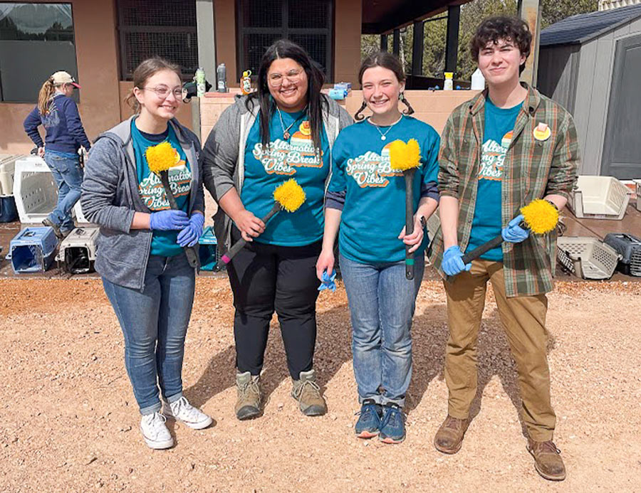 four students wear branded shirts, blue latex gloves and hold bright yellow bristle scrub brushes while taking a group photo at an outdoor event
