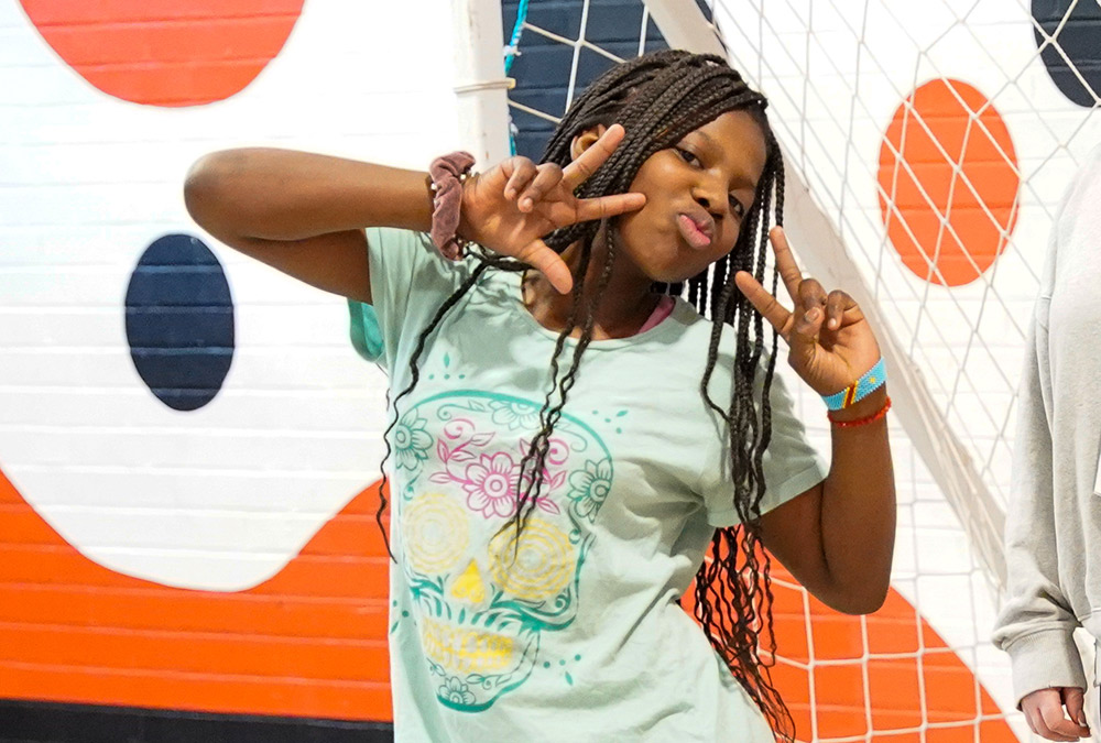 a young middle school girl with long braids poses and holds her hands in peace signs for a photo while playing soccer at an Amiko Youth Program event