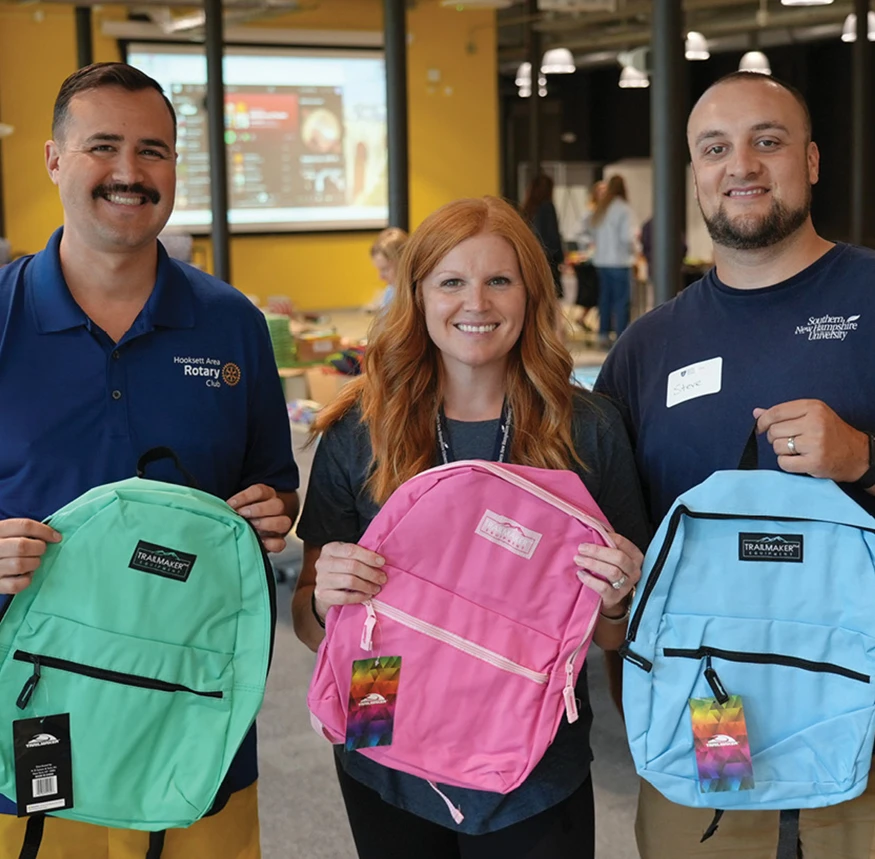 Three volunteers pose indoors holding colorful backpacks during a community donation or school supply drive event.