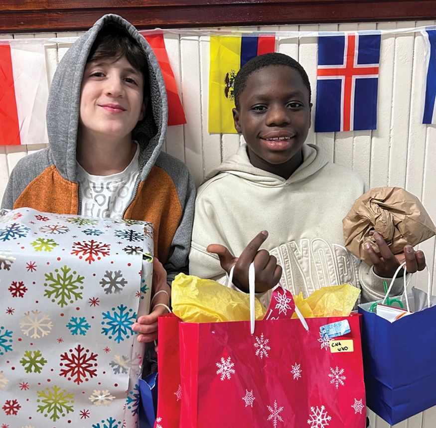 Two children hold wrapped gifts and holiday gift bags while standing in front of a wall decorated with international flags.