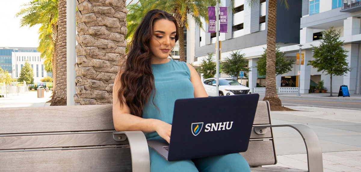 A wide-angle shot of a woman in a turquoise dress or blouse sitting on a park bench outdoors, working on a laptop featuring a Southern New Hampshire University (SNHU) logo, with modern campus buildings and palm trees in the background