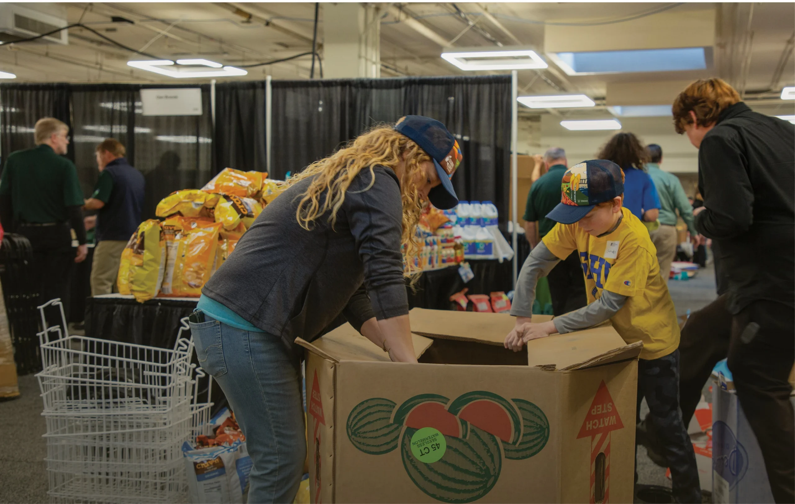 Volunteers pack supplies into large boxes at an indoor service event, with food and donation items visible on nearby tables.