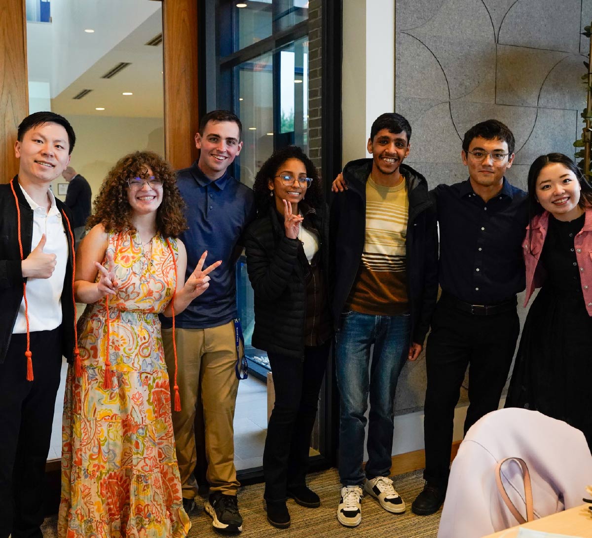 A diverse group of seven young adults stands together indoors, smiling and posing for a group photo.