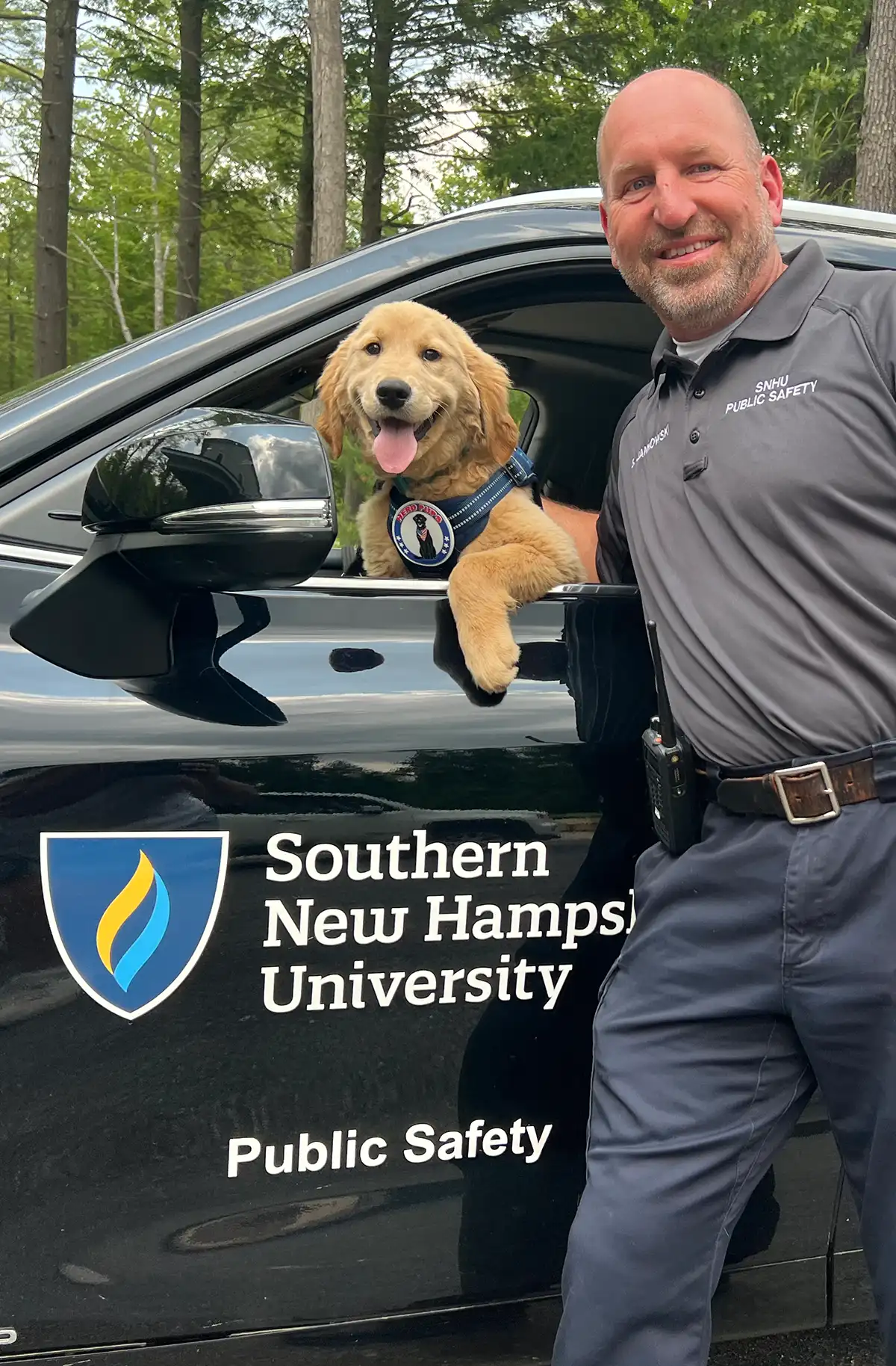 An SNHU Public Safety officer standing next to a patrol vehicle with a golden retriever service dog puppy wearing a "Hero Pups" vest leaning out of the window.