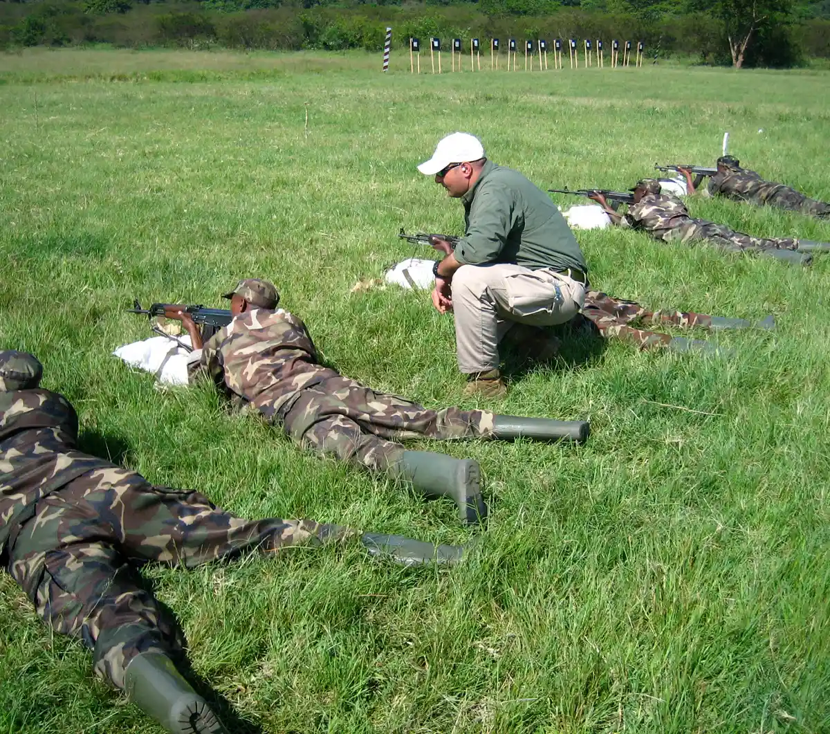 A photograph of a military training exercise in a grassy field, featuring a civilian instructor in a white cap and olive shirt supervising soldiers in camouflage uniforms as they practice marksmanship from a prone position